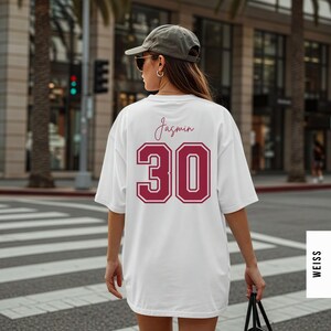 May include: A white oversized t-shirt featuring the name Jasmin in a dark red script font above the large dark red number 30 with a white outline. The shirt is worn with a grey baseball cap and dark sunglasses, viewed from the back.