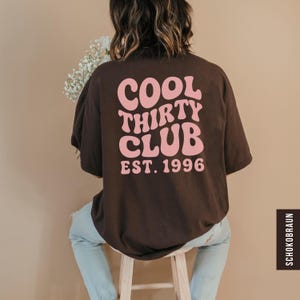 May include: A brown t-shirt with the text "COOL THIRTY CLUB EST. 1996" in pink, a vintage font. The shirt is worn by a person sitting on a wooden stool. The person is wearing light blue jeans.
