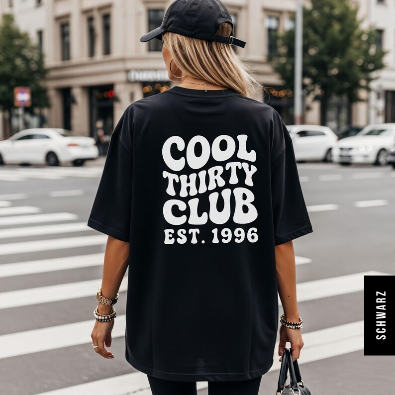 Puede incluir: Una camiseta negra de manga corta con texto blanco en la espalda que dice COOL THIRTY CLUB EST. 1996 en una fuente ondulada de estilo retro. La camiseta tiene un corte holgado y extragrande, y se lleva con una gorra negra y pulseras.
