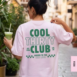 May include: A light pink short-sleeve t-shirt featuring dark green text on the back that reads COOL THIRTY CLUB, EST 1996, LVL 30, and CHEERS TO THE NEXT LEVEL, accompanied by a small checkered pattern graphic at the bottom of the design.