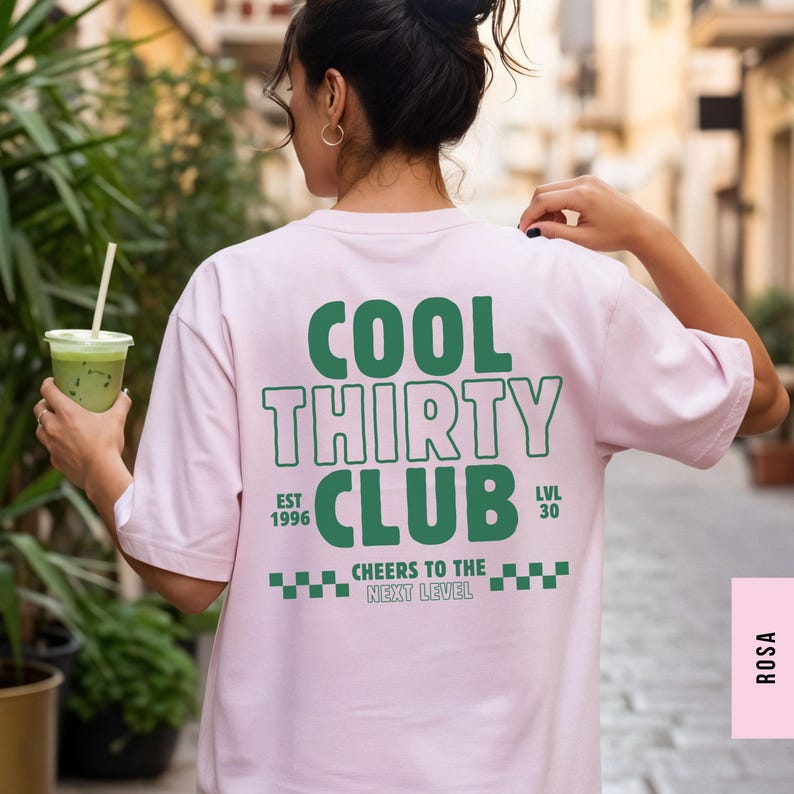 May include: A light pink short-sleeve t-shirt featuring dark green text on the back that reads COOL THIRTY CLUB, EST 1996, LVL 30, and CHEERS TO THE NEXT LEVEL, accompanied by a small checkered pattern graphic at the bottom of the design.