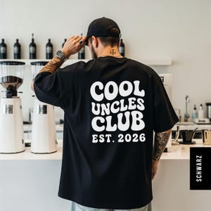 K&ouml;nnte beinhalten: Schwarzes T-Shirt mit dem wei&szlig;en Text "COOL UNCLES CLUB EST. 2026" in einem Retro-Schriftstil. Das Shirt ist &uuml;bergro&szlig; und hat kurze &Auml;rmel. Die Person tr&auml;gt das Shirt in einem Caf&eacute;.