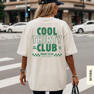 May include: An oversized cream-colored t-shirt featuring dark green text on the back that reads COOL THIRTY CLUB, EST 1996, LVL 30, and CHEERS TO THE NEXT LEVEL, accompanied by a checkered pattern graphic design.