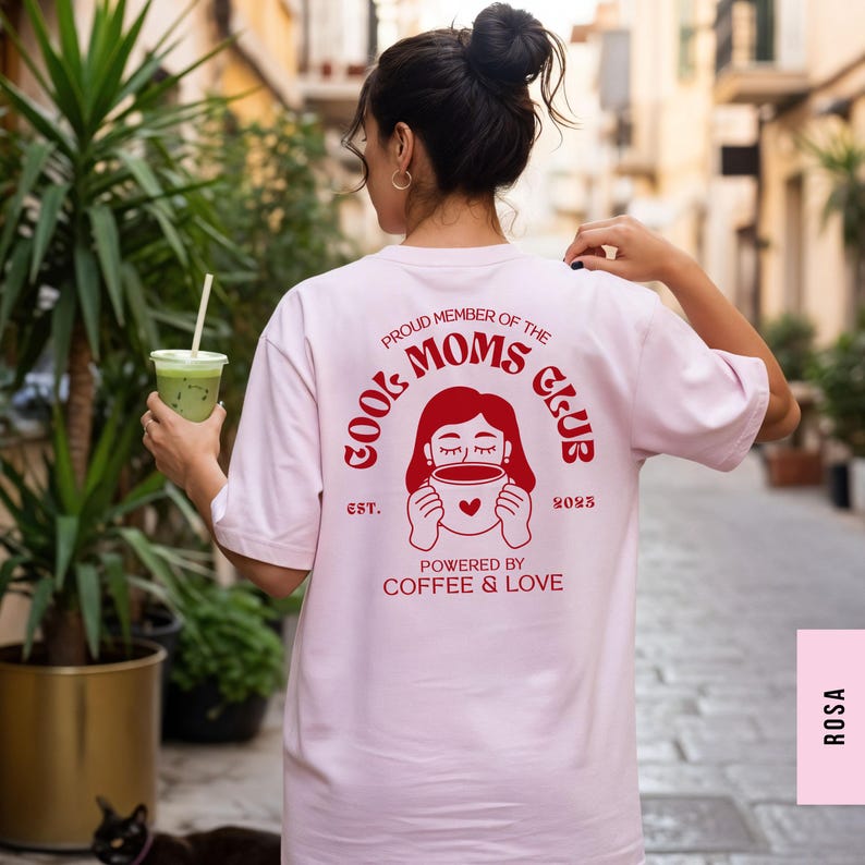 May include: A light pink t-shirt with the text "COOL MOMS CLUB" in red, along with a graphic of a woman holding a coffee cup. The text "PROUD MEMBER OF THE" and "POWERED BY COFFEE & LOVE" are also printed on the back.