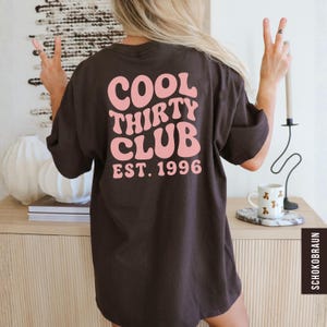May include: A dark brown t-shirt with the text "COOL THIRTY CLUB EST. 1996" in pink, retro-style lettering. The shirt is a casual, oversized fit. The image shows the back of the shirt.