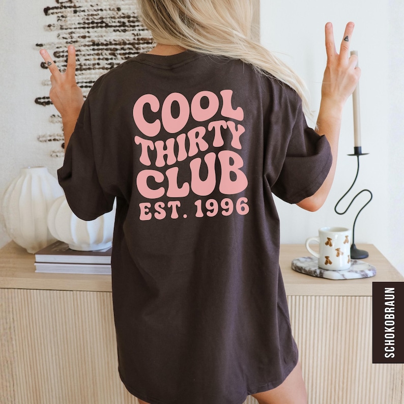 May include: A dark brown t-shirt with the text "COOL THIRTY CLUB EST. 1996" in pink, retro-style lettering. The shirt is a casual, oversized fit. The image shows the back of the shirt.