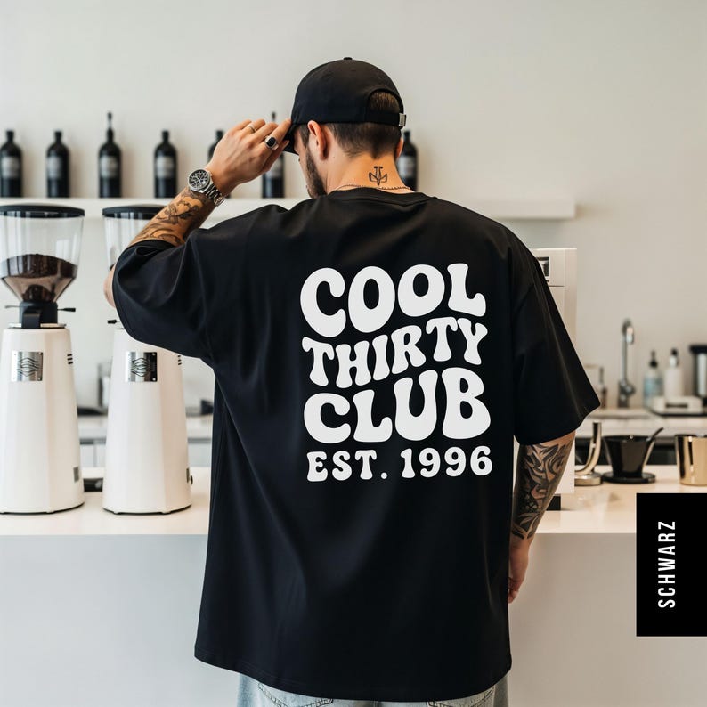 Puede incluir: Una camiseta negra de manga corta con texto blanco en la espalda que dice COOL THIRTY CLUB EST. 1996 en una fuente ondulada de estilo retro. La camiseta tiene un corte holgado y relajado, y se lleva con una gorra negra y pantalones vaqueros.