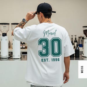 Op de afbeelding: Wit T-shirt met de naam "Marcel" en het nummer "30" in groen, samen met "EST. 1996" op de achterkant gedrukt. De persoon op de foto draagt een zwarte baseball cap.