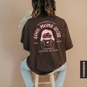 Könnte beinhalten: Dunkelbraunes T-Shirt mit rosa Grafik, die "Cool Moms Club" mit einer Frau, die eine Kaffeetasse hält, zeigt. Der Text "Powered by Coffee & Love" steht darunter. Das Shirt hat eine lockere Passform.