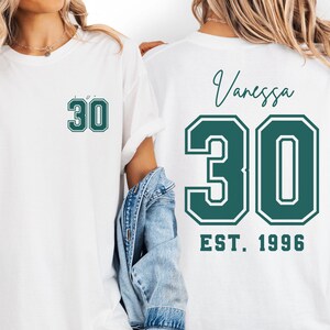 Op de afbeelding: Witte T-shirts met het nummer "30" in een groen en wit ontwerp. E&eacute;n shirt heeft het nummer op de voorkant, en de andere heeft het nummer, de naam "Vanessa" en "EST. 1996" op de achterkant. De shirts zijn casual en waarschijnlijk voor een verjaardag.