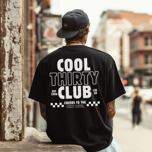 May include: A black short-sleeve t-shirt featuring white text on the back that reads COOL THIRTY CLUB, EST 1996, LVL 30, and CHEERS TO THE NEXT LEVEL, accompanied by a checkered pattern. The shirt is paired with a dark gray baseball cap with a logo.
