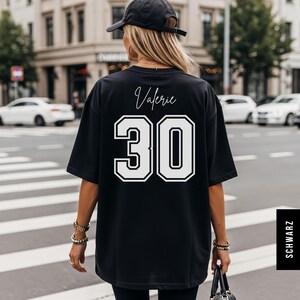 May include: A black oversized t-shirt featuring the name Valerie in white script font above the large white number 30 in a varsity style block typeface. The shirt is worn with a black baseball cap, and the back of the garment is displayed.
