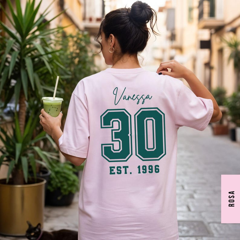 Op de afbeelding: Lichtroze T-shirt met de naam "Vanessa" in cursief boven het nummer "30" in groene letters. Daaronder staat "EST. 1996". Het T-shirt heeft een casual stijl.