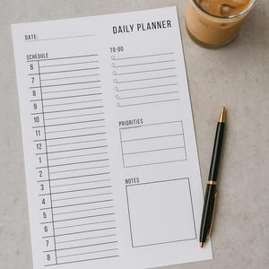 Op de afbeelding: Een witte dagplanner met de tekst "DAILY PLANNER" en secties voor schema, taken, prioriteiten en notities. Een zwarte en gouden pen en een glas ijskoffie liggen naast de planner.