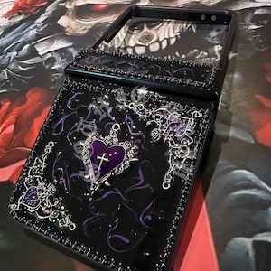 May include: Black phone case with a flip-up mirror, decorated with a purple heart pendant and cross. The case features silver filigree, purple swirls, and black bead trim. The design has a gothic aesthetic.