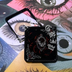 May include: Black phone case with a decorative design featuring a bat, ornate silver accents, and red swirls. The case has a glossy finish and a black frame. The background features eye illustrations.