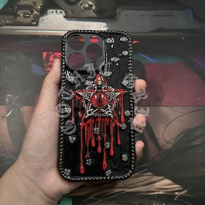 Könnte beinhalten: Schwarze Handyhülle mit einem dekorativen Sterndesign. Der Stern hat ein rotes Auge in der Mitte und ist von silbernen Akzenten umgeben. Rote Tropfen gehen vom Stern aus. Die Hülle hat einen schwarzen Rand mit silbernen Nieten.
