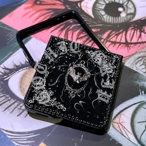 May include: Black phone case with a decorative design featuring a bat, stars, and ornate lettering. The case has a glossy finish and a black frame. The design is in silver and white. The case is on a colorful background.
