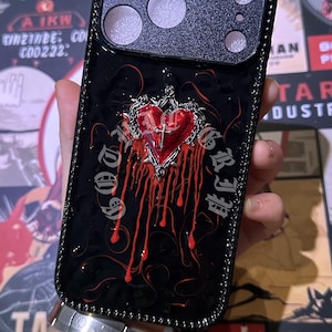Op de afbeelding: Zwarte telefoonhoes met een decoratief ontwerp met een rood hart, een kruis en een druipend bloedeffect. De hoes heeft een zwarte rand en een gestructureerd camera-gedeelte.