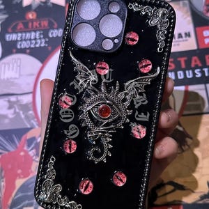 Peut inclure: Coque de téléphone noire avec un motif de dragon. La coque présente un dragon argenté avec des yeux et des accents en pierres précieuses rouges. Des détails supplémentaires d'yeux en pierres précieuses rouges sont répartis sur la surface noire brillante.