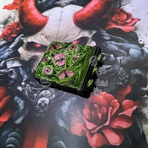 May include: A square, green and black object with multiple pink and white eyes on the top surface. The object is set against a backdrop of a skull with red horns, roses, and a sword.