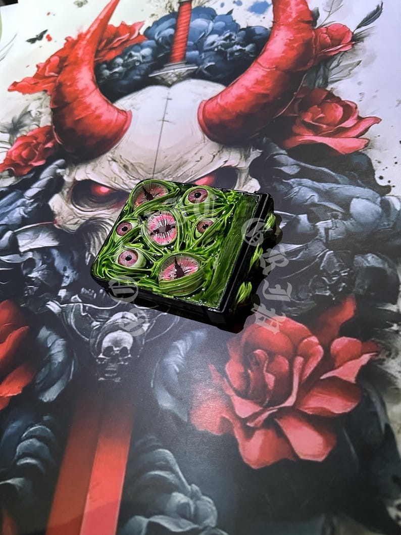 May include: A square, green and black object with multiple pink and white eyes on the top surface. The object is set against a backdrop of a skull with red horns, roses, and a sword.