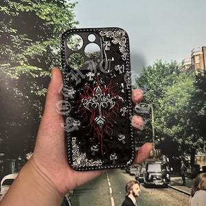 Peut inclure: Coque de téléphone noire avec un motif de croix décoratif. L'étui présente une bordure et des accents argentés, avec un motif de croix rouge et argenté au centre. L'étui comporte des découpes pour l'appareil photo et les boutons.