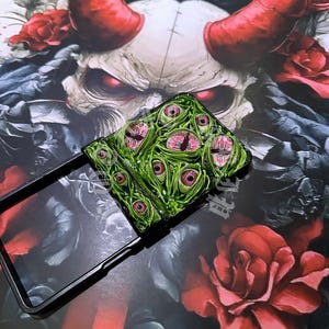 May include: A black phone case with a green and pink eye design. The case is open, revealing the phone's screen. The background features a skull with red horns and roses, creating a dark, gothic aesthetic.