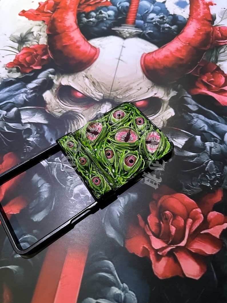 May include: A black phone case with a green and pink eye design. The case is open, revealing the phone's screen. The background features a skull with red horns and roses, creating a dark, gothic aesthetic.