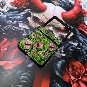 May include: A black phone case featuring a green, textured design with multiple pink and red eyes. The case is set against a backdrop of a demonic illustration with red horns and roses.
