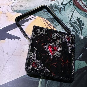 Puede incluir: Bolso de mano negro con un diseño decorativo que presenta un corazón rojo y adornos plateados. El bolso tiene un asa y está adornado con texto. La estética general es gótica.