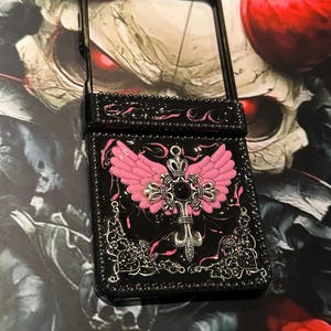 Könnte beinhalten: Eine schwarze, rechteckige Handyhülle mit glänzender Oberfläche und rosa Marmorierung. In der Mitte befindet sich ein silberfarbenes Kreuz-Emblem mit rosa Flügeln und einem schwarzen Stein. Dekorative silberne Filigran-Akzente zieren die unteren Ecken.