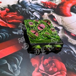 May include: A rectangular, green, and black decorative box featuring multiple eye designs with pink irises. The box is set against a backdrop of red roses and a dark, artistic background.