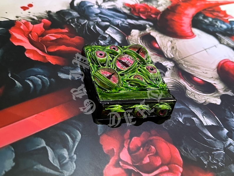 May include: A rectangular, green, and black decorative box featuring multiple eye designs with pink irises. The box is set against a backdrop of red roses and a dark, artistic background.