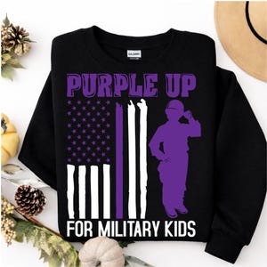 Puede incluir: Sudadera negra de cuello redondo con el texto "PURPLE UP" en morado. El dise&ntilde;o incluye una silueta morada de una persona uniformada y un dise&ntilde;o de bandera morada y blanca. El texto "FOR MILITARY KIDS" est&aacute; debajo.