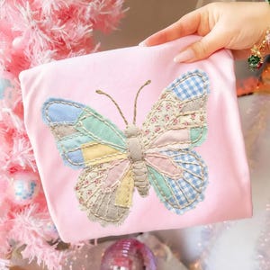 Może przedstawiać: Jasnoróżowa koszulka z dużą, aplikowaną motylem. Skrzydła motyla są wykonane z patchworku, z sekcjami niebieskiej kratki vichy, kwiatowych nadruków i pastelowych tkanin. Ciało motyla jest w kolorze beżowym, a czułki to cienkie, brązowe nici.