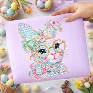 Pode incluir: Uma almofada lavanda com um design de coelho da Páscoa. O coelho tem óculos com armação dourada e um padrão floral. O design inclui um laço e flores. Ovos de Páscoa e pintinhos no fundo.