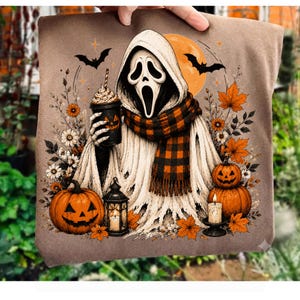 May include: A brown t-shirt featuring a graphic of a hooded ghost figure wearing an orange and black plaid scarf, holding a coffee cup with a jack-o'-lantern face. The design includes two pumpkins, a lantern, a candle, bats, and autumn leaves.