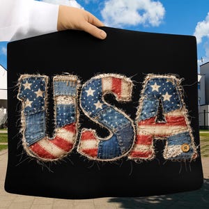Op de afbeelding: Zwarte achtergrond met het woord "USA" in een patchwork-ontwerp. De letters zijn gemaakt van denim en stof met rood, wit en blauwe patronen, inclusief sterren en strepen. De randen zijn omrand met een draad van natuurlijke kleur.