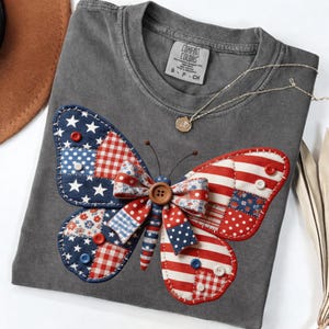 May include: A gray Comfort Colors t-shirt featuring a patriotic butterfly design. The butterfly wings are made of red, white, and blue fabric patches, with button accents. A gold necklace is draped on the shirt.