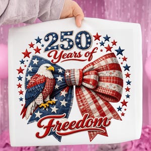 Op de afbeelding: Een wit vierkant kussen met een patriottisch ontwerp. Het ontwerp toont een strik met rood-witte ruit- en bloempatronen, een Amerikaanse vlag en een adelaar. De tekst luidt "250 Years of Freedom".