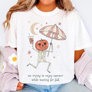 May include: A white t-shirt featuring a graphic of a skeleton with a pumpkin head holding a drink and a striped umbrella. The design includes a crescent moon and stars, with the text "me trying to enjoy summer while waiting for fall."