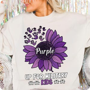 Puede incluir: Sudadera color crema con un gran girasol morado y la palabra "Purple" en el centro. Encima de la flor hay huellas de manos moradas. El texto "UP FOR MILITARY KIDS" est&aacute; debajo.
