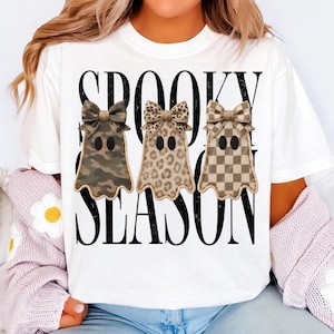 May include: A white t-shirt featuring the black text SPOOKY SEASON. The design includes three ghost shapes, each topped with a bow. The first ghost has a camouflage pattern, the second has a leopard print, and the third has a tan checkerboard pattern.