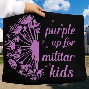 Puede incluir: Bolso tote negro con un dise&ntilde;o de diente de le&oacute;n morado y el texto "purple up for militar kids". El bolso se sostiene contra un cielo azul. El dise&ntilde;o es en tonos morados.