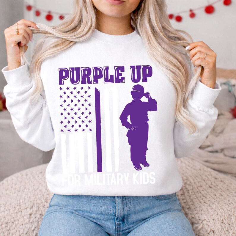Puede incluir: Sudadera blanca de cuello redondo con el texto "PURPLE UP" en morado sobre una silueta morada de una persona saludando y un dise&ntilde;o de bandera estadounidense morada. El texto "FOR MILITARY KIDS" est&aacute; debajo.