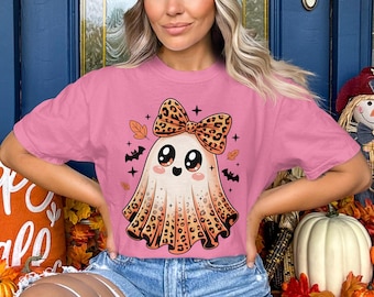 May include: Pink t-shirt featuring a cute ghost with a leopard print bow and skirt. The ghost has large eyes and is surrounded by bats, leaves, and stars. A fun and festive design for Halloween.