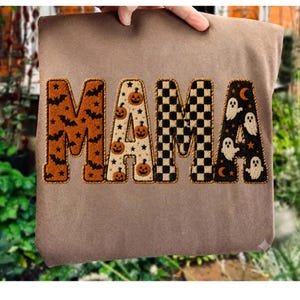 May include: A brown sweatshirt featuring the word MAMA in large block letters. Each letter has a distinct pattern: the M has orange bats, the A has orange pumpkins, the M has a black and white checkerboard, and the A has white ghosts.