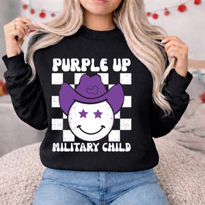 Puede incluir: Sudadera negra con el texto "PURPLE UP MILITARY CHILD". El dise&ntilde;o presenta una carita sonriente con un sombrero de vaquero morado con dos ojos de estrella, sobre un fondo a cuadros blanco y negro.