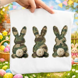 Può includere: Federa bianca con tre sagome di conigli. I conigli hanno corpi in tessuto mimetico e soffici code bianche. Il design crea un tema pasquale. La federa è tenuta in mano.